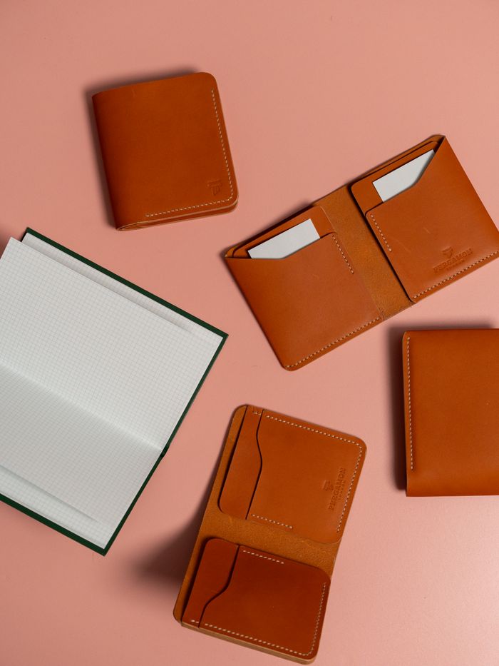 Flat lay of stylish tan leather wallets and notebook on a pastel pink backdrop.