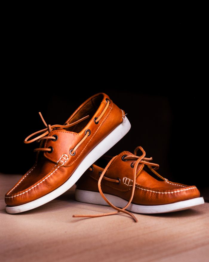 Close-up of brown leather boat shoes with white soles on a wooden floor against a black background.